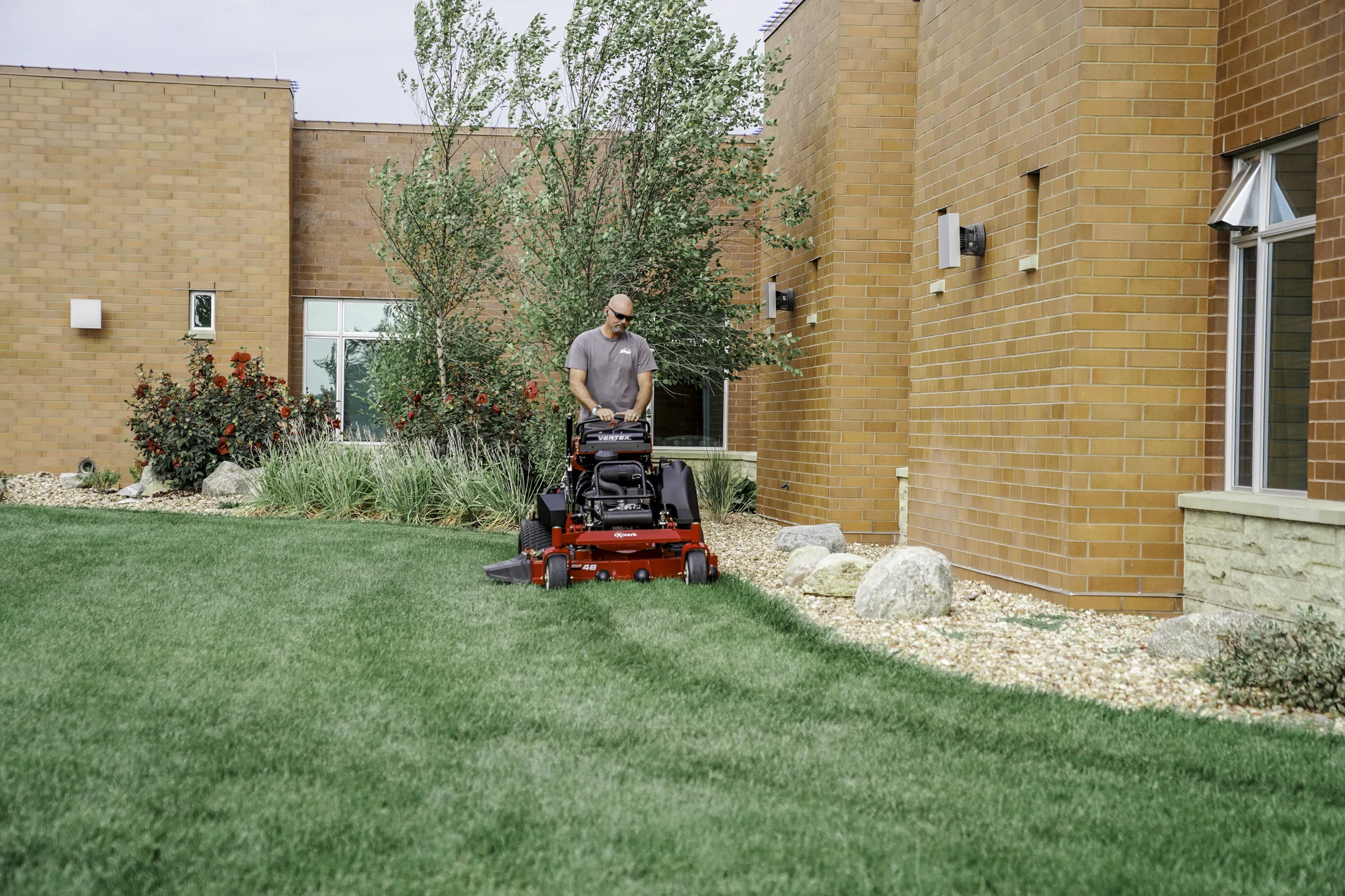 Exmark Vertex S Series Stand-On Lawn Mower VXS740EKC52400 Exmark Vertex S Series Stand On Lawn Mower Vxs740Ekc52400 Man Mowing Near Building product image