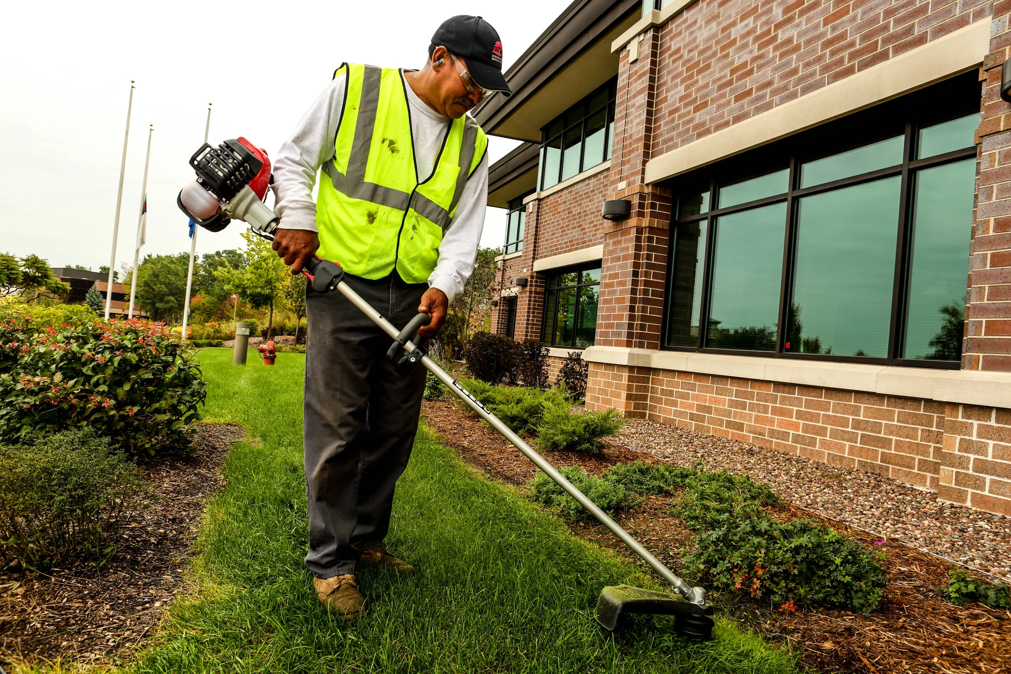 Shindaiwa Trimmer T262 Shindaiwa T Series Trimmer T262 Lifestyle Wide Shot Right Side Man Trimming Grass Side Of Building product image